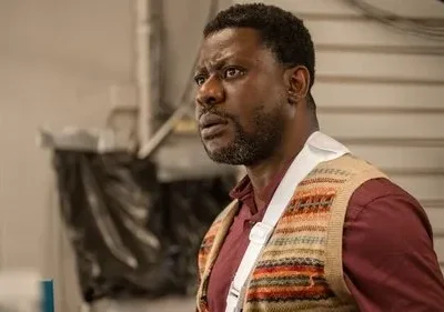 Homem negro de camisa vinho, colete estampado e tipoia, olha com apreensão. Fundo claro, ambiente interno simples.