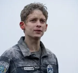 Jovem com cabelo cacheado, rosto sujo e arranhado, veste uniforme de polícia empoeirado. Fundo cinza. Clima sombrio e exausto.