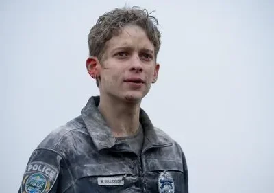 Jovem com cabelo cacheado, rosto sujo e arranhado, veste uniforme de polícia empoeirado. Fundo cinza. Clima sombrio e exausto.