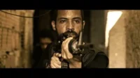 Homem barbudo e suado, usando um terço, mira um rifle num beco escuro. Clima intenso e sépia.
