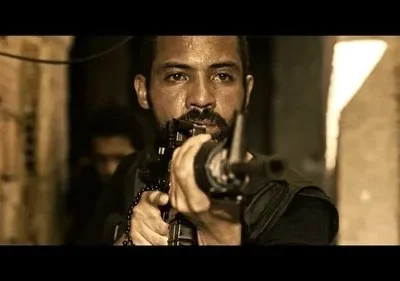 Homem barbudo e suado, usando um terço, mira um rifle num beco escuro. Clima intenso e sépia.