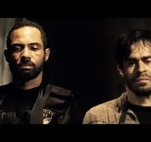 Dois homens com expressões sérias em um ambiente escuro. Um usa colete tático preto, o outro uma camisa bege. Clima sombrio.