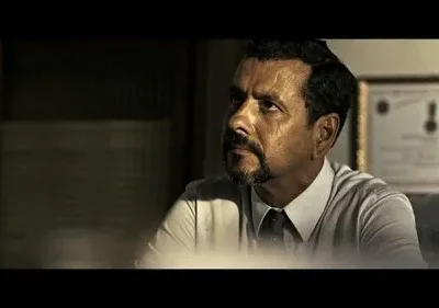 Um homem sério de barba, camisa e gravata, olha pensativo para cima. Cenário de escritório escuro com diploma. Clima tenso e dramático.