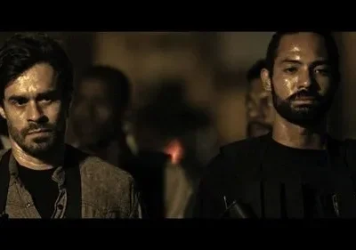 Dois homens, um de colete tático, outro de camisa, com rostos suados e sérios. Cenário noturno escuro e tenso.