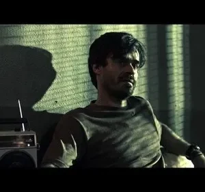 Um homem barbudo e pensativo sentado em um quarto escuro. Sombras de persianas caem sobre a parede e um rádio vintage ao lado. Clima sombrio.