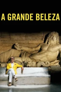 Homem de óculos e casaco amarelo brilhante senta-se à frente de uma estátua clássica de mármore de um homem barbudo reclinado. Cenário grandioso.