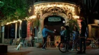 Noite. Grupo de jovens com bicicletas e um cão em frente a um prédio ricamente decorado e iluminado com FOREVER. Clima urbano.