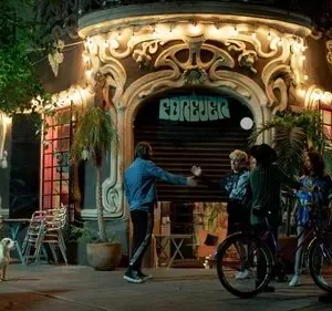Noite. Grupo de jovens com bicicletas e um cão em frente a um prédio ricamente decorado e iluminado com FOREVER. Clima urbano.