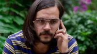 Homem barbudo de cabelo longo, óculos e camisa listrada, fala ao celular com expressão séria. Fundo de folhagem verde.
