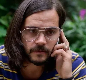 Homem barbudo de cabelo longo, óculos e camisa listrada, fala ao celular com expressão séria. Fundo de folhagem verde.