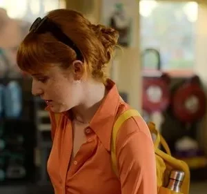 Jovem ruiva com óculos na cabeça, camisa laranja e mochila amarela, parece pensativa em um ambiente interno casual.