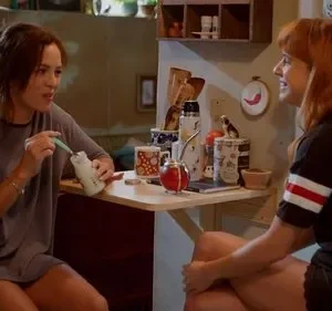 Duas mulheres conversam em um ambiente doméstico. Uma, com mamadeira, e a outra, de cabelo ruivo, sorri, transmitindo clima casual.
