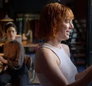 Mulher ruiva sorridente examina roupas. Ao fundo, mulher séria senta à mesa num ateliê. Clima de trabalho e foco.