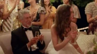 Homem de smoking e mulher de branco sorriem em festa elegante. Convidados celebram brindando sob luzes festivas.
