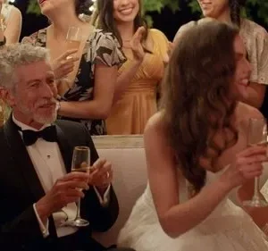Homem de smoking e mulher de branco sorriem em festa elegante. Convidados celebram brindando sob luzes festivas.