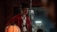 Mulher com coques e batom vermelho, de camisa vermelha aberta, canta em microfone vintage num estúdio escuro. Clima musical intenso.