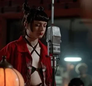 Mulher com coques e batom vermelho, de camisa vermelha aberta, canta em microfone vintage num estúdio escuro. Clima musical intenso.