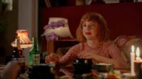 Mulher ruiva de batom vermelho e camisa rosa olha atenta à mesa com comida, velas e lâmpadas. Clima íntimo.