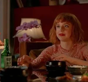 Mulher ruiva de batom vermelho e camisa rosa olha atenta à mesa com comida, velas e lâmpadas. Clima íntimo.