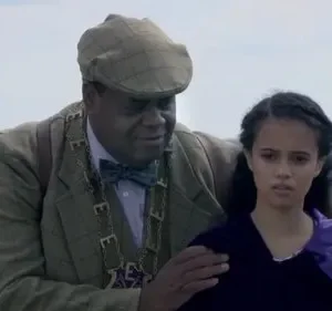 Homem de casaco tweed e gorro segura o ombro de jovem de vestido roxo com expressão séria. Fundo: colinas e flora púrpura.