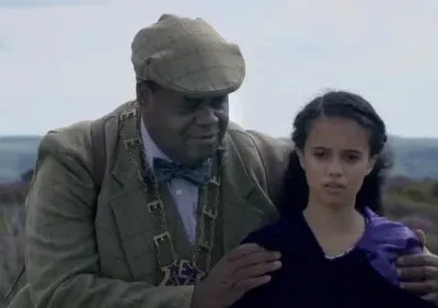 Homem de casaco tweed e gorro segura o ombro de jovem de vestido roxo com expressão séria. Fundo: colinas e flora púrpura.