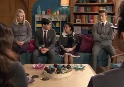 Cinco jovens de uniforme sentados em um sofá, alguns sérios, um adulto de costas à direita. Uma sala com estantes de livros.