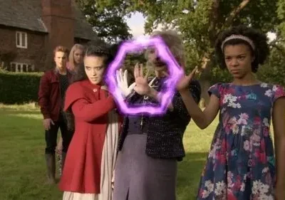 Mulher mais velha com hexágono mágico roxo nas mãos, ladeada por jovens em campo gramado. Casa de tijolos ao fundo.