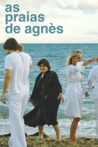 Três pessoas descalças na beira do mar,incluindo uma mulher mais velha sorrindo. Céu nublado,atmosfera alegre.