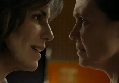 Duas mulheres de perfil, rostos muito próximos. Uma sorri sutilmente, a outra olha com seriedade. Atmosfera íntima e tensa.