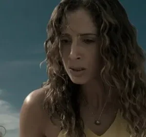 Mulher com cabelos cacheados olha com expressão aflita para criança. Céu azul e nuvens ao fundo.