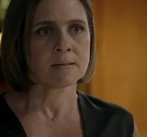 Close-up de mulher com cabelo curto, olhar apreensivo, vestindo blusa escura. Fundo desfocado de luz amarela e parede marrom.