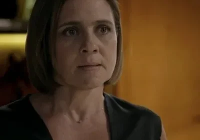 Close-up de mulher com cabelo curto, olhar apreensivo, vestindo blusa escura. Fundo desfocado de luz amarela e parede marrom.