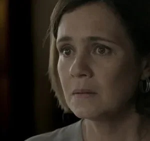 Mulher de meia-idade, cabelo curto, com olhar preocupado e olhos arregalados. Em um ambiente escuro e tenso.