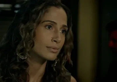 Mulher de cabelos cacheados olha seriamente para o lado. Um homem sombrio e barbudo desfocado está ao fundo. Clima misterioso.