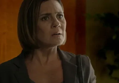 Mulher de cabelo castanho e blazer escuro exibe expressão de choque ou preocupação intensa. Fundo interno desfocado com tons quentes.