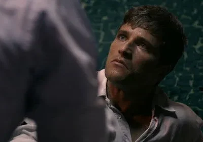 Homem com camisa clara e suja olha para cima com intensidade. Parte de outra figura em primeiro plano. Fundo escuro rachado.