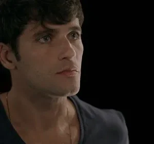 Jovem homem moreno de olhos claros olha pensativo para o lado, contra um fundo escuro. Clima dramático e introspectivo.