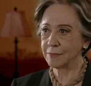 Retrato de mulher idosa com cabelos grisalhos e colar dourado, em ambiente interno. Ela tem expressão digna e observadora.