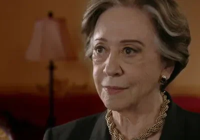 Retrato de mulher idosa com cabelos grisalhos e colar dourado, em ambiente interno. Ela tem expressão digna e observadora.