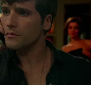 Homem sério fala ao telefone no primeiro plano. Uma mulher desfocada, de ombros nus, o observa no fundo escuro. Clima de mistério.