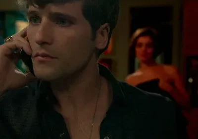 Homem sério fala ao telefone no primeiro plano. Uma mulher desfocada, de ombros nus, o observa no fundo escuro. Clima de mistério.