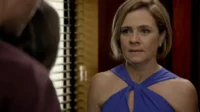 Close-up:Mulher loira em vestido azul com expressão séria olha para figura desfocada à esquerda. Fundo com persianas e tons escuros.