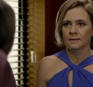 Close-up: Mulher loira em vestido azul com expressão séria olha para figura desfocada à esquerda. Fundo com persianas e tons escuros.