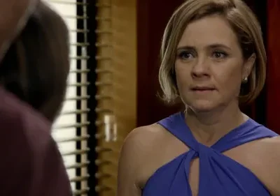 Close-up: Mulher loira em vestido azul com expressão séria olha para figura desfocada à esquerda. Fundo com persianas e tons escuros.