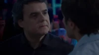 Dois homens conversam em um ambiente escuro. Um homem mais velho e sério olha para um homem mais jovem,visto de costas. Luzes coloridas ao fundo.