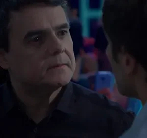 Dois homens conversam em um ambiente escuro. Um homem mais velho e sério olha para um homem mais jovem, visto de costas. Luzes coloridas ao fundo.