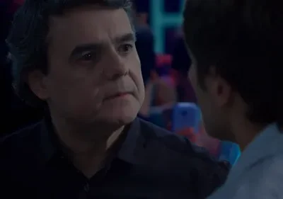 Dois homens conversam em um ambiente escuro. Um homem mais velho e sério olha para um homem mais jovem, visto de costas. Luzes coloridas ao fundo.