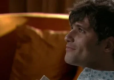 Homem jovem de cabelo escuro, em camisa clara pontilhada, sorri olhando para cima em ambiente com fundo quente e desfocado.