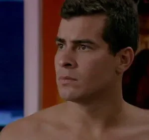 Homem sério e sem camisa olha para o lado. Fundo em tons de azul e vermelho sugere cenário moderno e tenso.