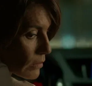 Close-up de mulher de cabelo castanho com olhar sério. Ela está em um carro escuro, à noite, com luzes de fundo. Clima misterioso e tenso.